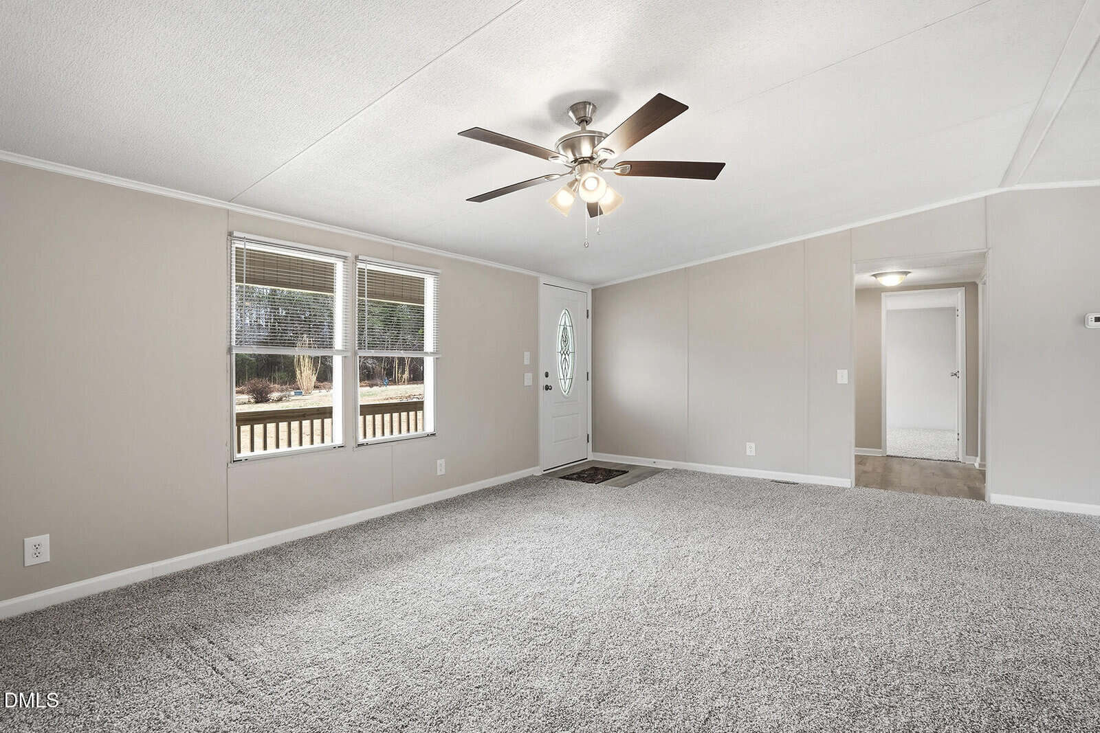 A neutral-toned living room with light beige walls, gray carpet, a ceiling fan with lights, two windows, a white front door with a glass oval, and an open doorway leading to another room.