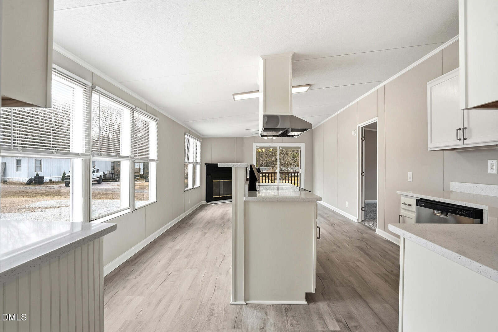 A modern kitchen with light wood flooring, white cabinets, an island with a range hood, large windows on the left, and a dishwasher on the right. Natural light fills the open space.
