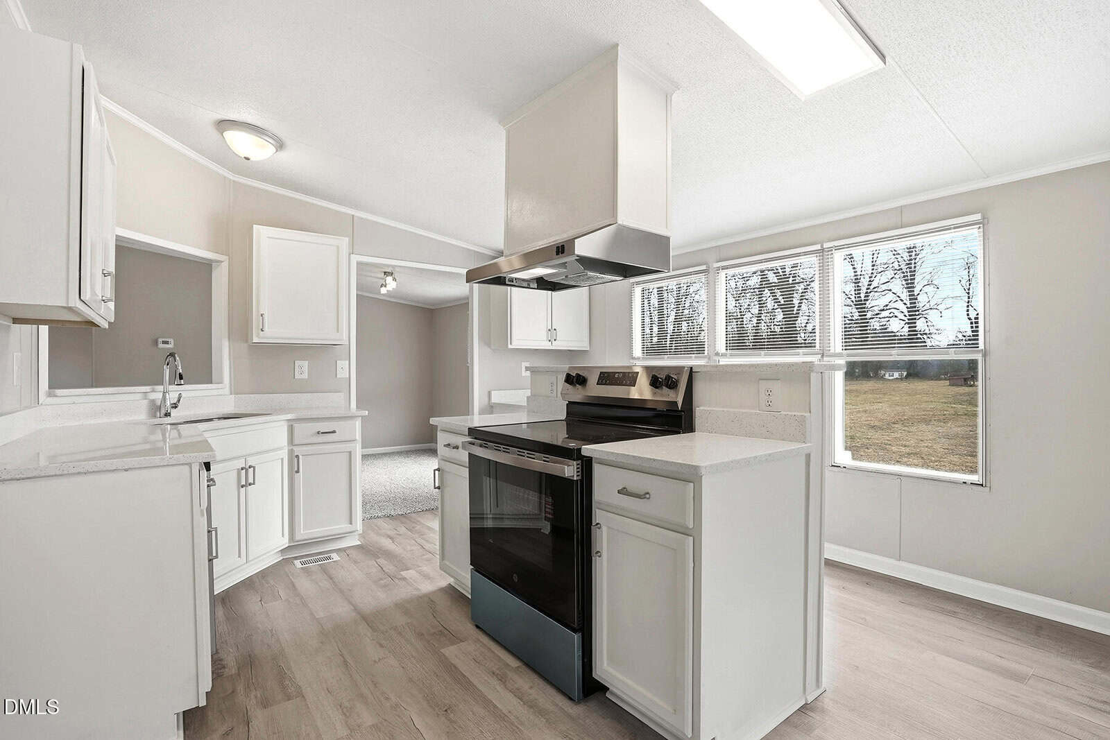 Modern kitchen with white cabinets, stainless steel stove, overhead vent, light wood floors, and large windows letting in natural light. Open layout connects to another room and shows a view of the outdoors.