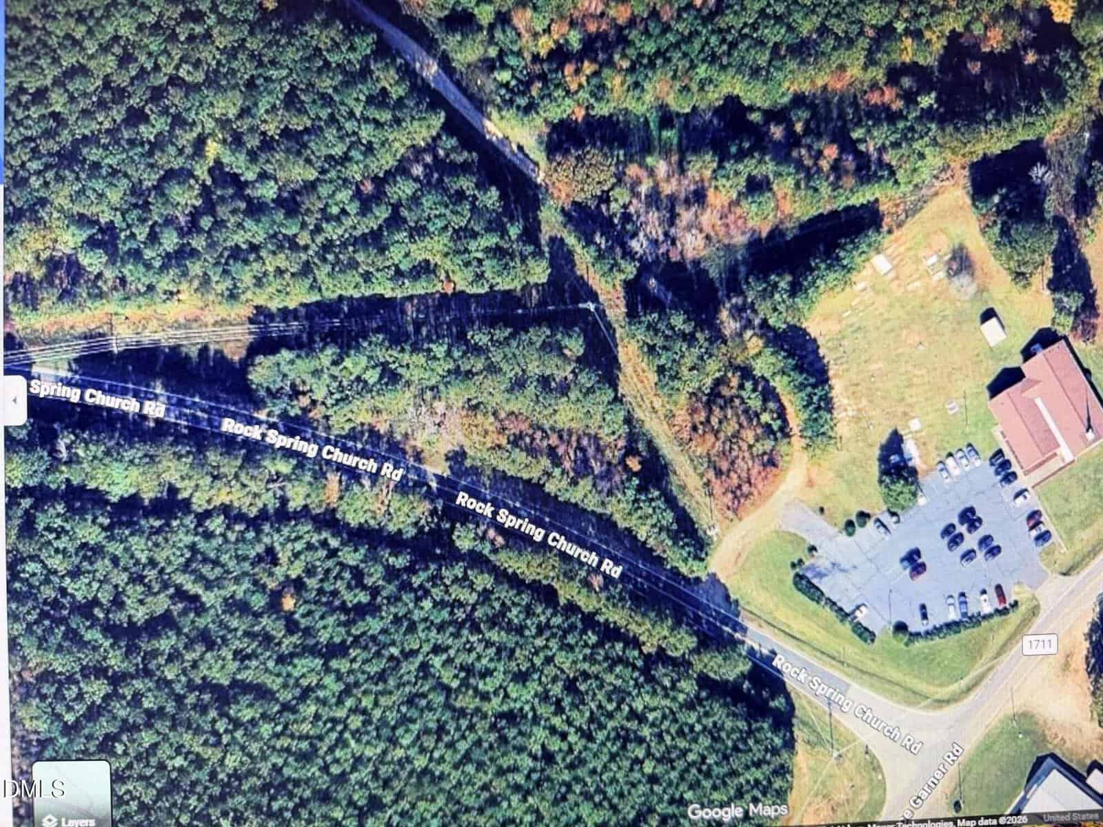 Aerial view of a wooded area intersected by Rock Spring Church Road, with a church building, parking lot with cars, and a small cemetery on the right side, next to a cleared grassy area.