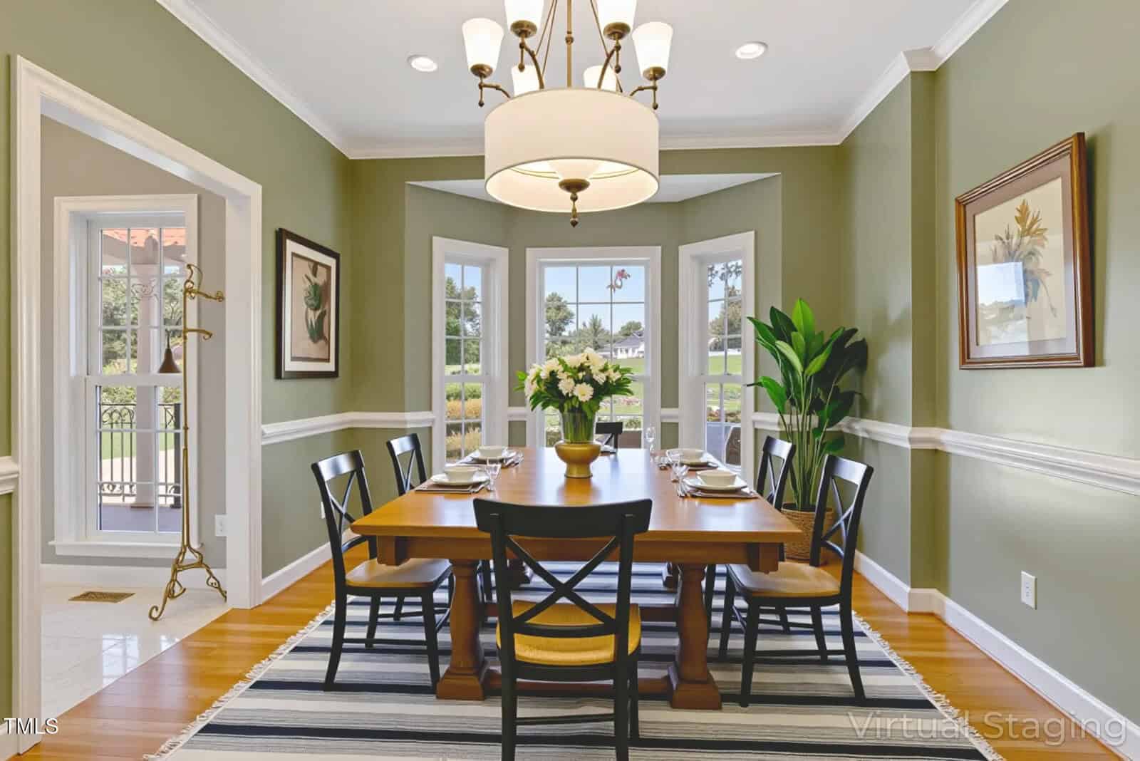 A dining room with green walls, a wooden table set for six with black chairs, a modern chandelier, large windows, a striped rug, a potted plant, framed artwork, and a side door to the outside.