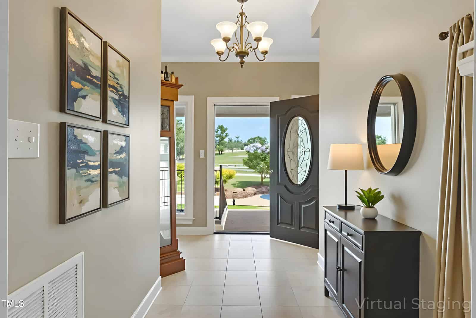 A view of a home entryway with beige walls, a tile floor, four abstract paintings on the left, a console table with a lamp and plant on the right, a round mirror above, and an open door showing a lawn outside.