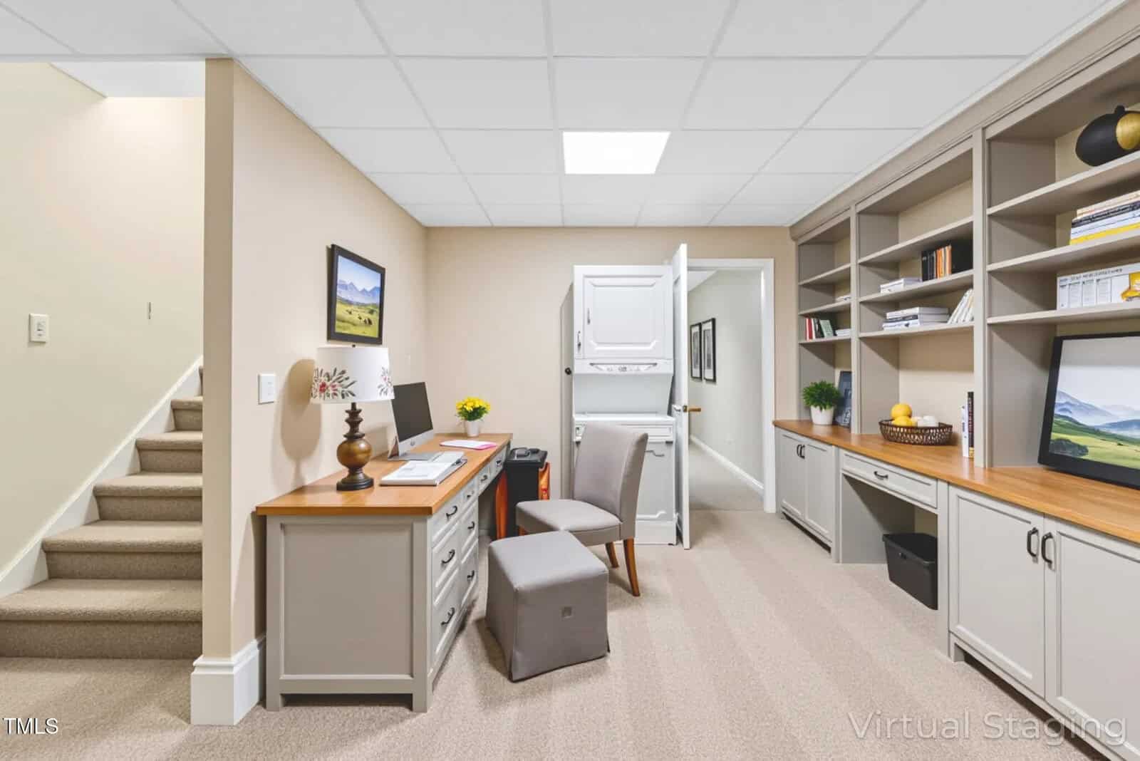 A home office features a wooden desk with papers and a lamp, a gray chair, built-in shelves with books and decor, a washer and dryer unit in the back, and a staircase leading upward on the left side.