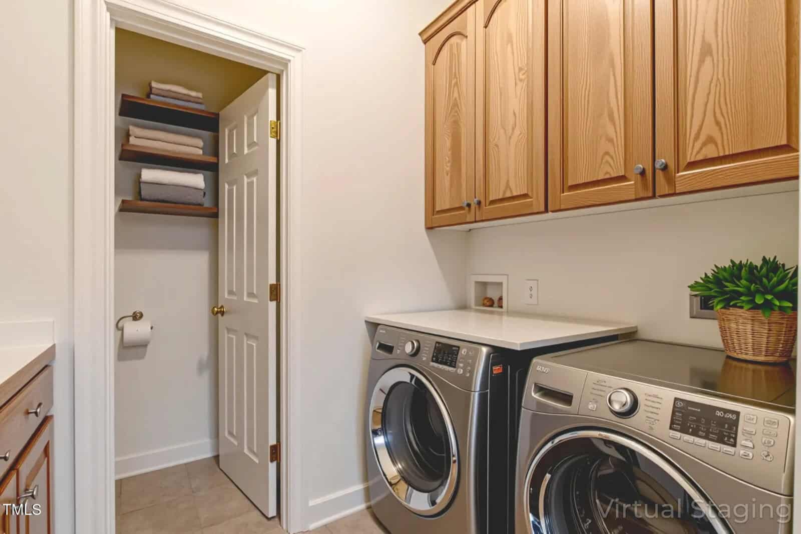 Modern laundry room with front-loading washer and dryer, wooden cabinets above, and a small closet with shelves holding folded towels and a toilet paper roll holder on the wall. A potted plant sits on the countertop.