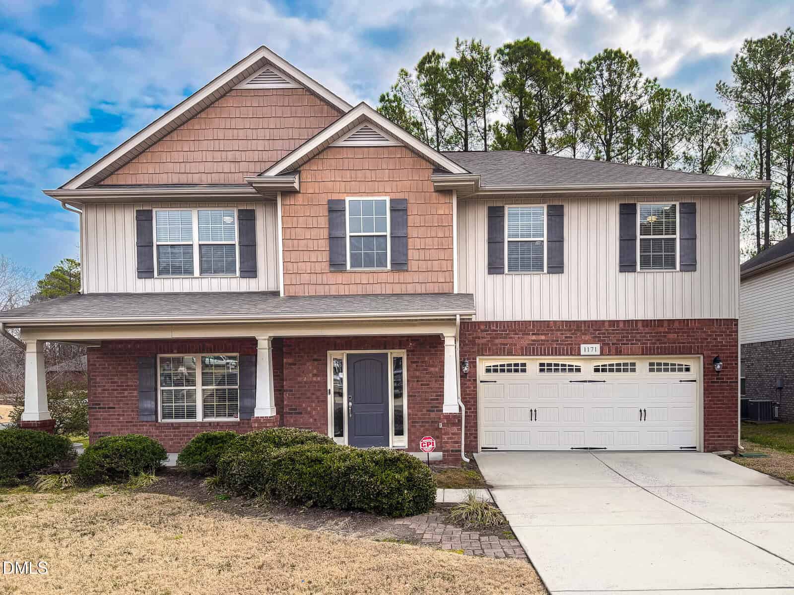 Charming two-story suburban house at 1171 Jackson Court in Creedmoor, featuring brick, shingle, and siding exterior, double garage, front porch, black shutters, and well-kept shrubs. Trees are visible in the background under a partly cloudy sky.