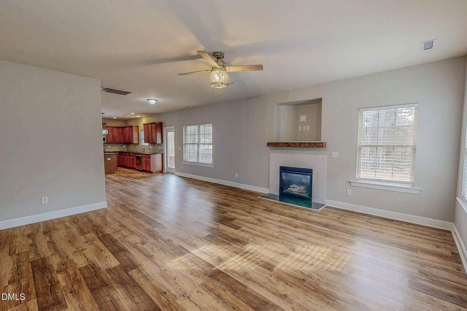 A spacious living area with wood flooring, beige walls, a ceiling fan, fireplace with a wooden mantel, and several windows. The living room opens to a kitchen with red cabinets and stainless steel appliances.