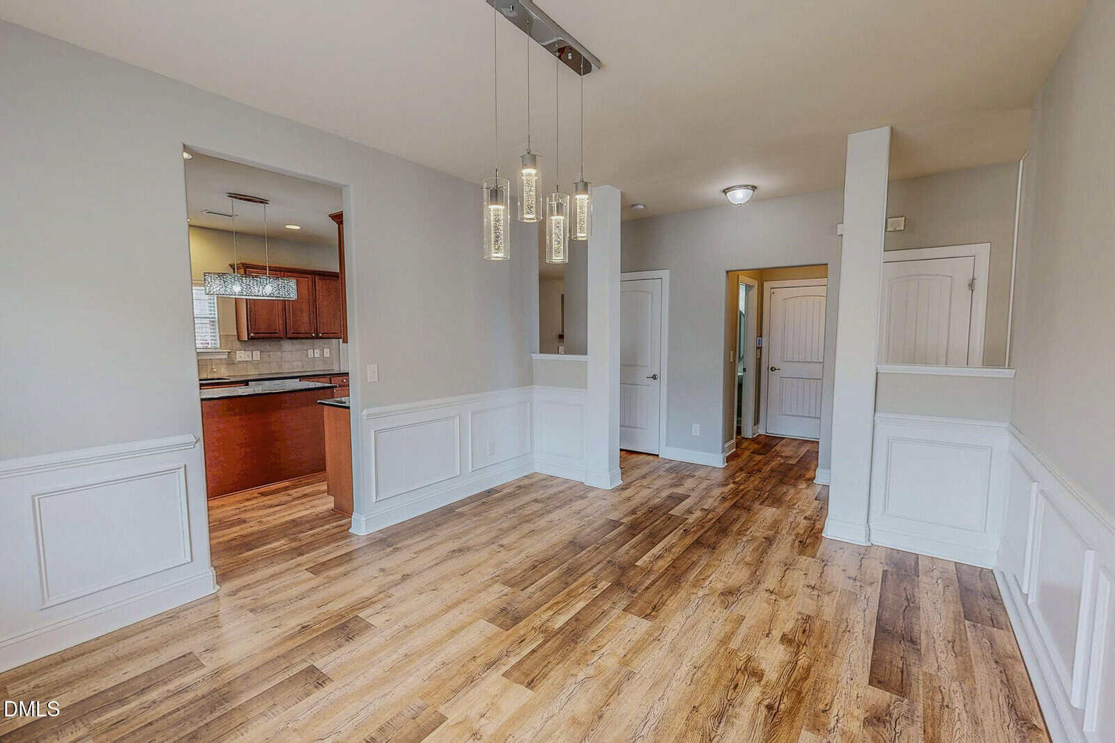 A dining area with wood flooring and white wainscoting, modern pendant lights overhead, and an open view into a kitchen with brown cabinets; a hallway leads to a door in the background.