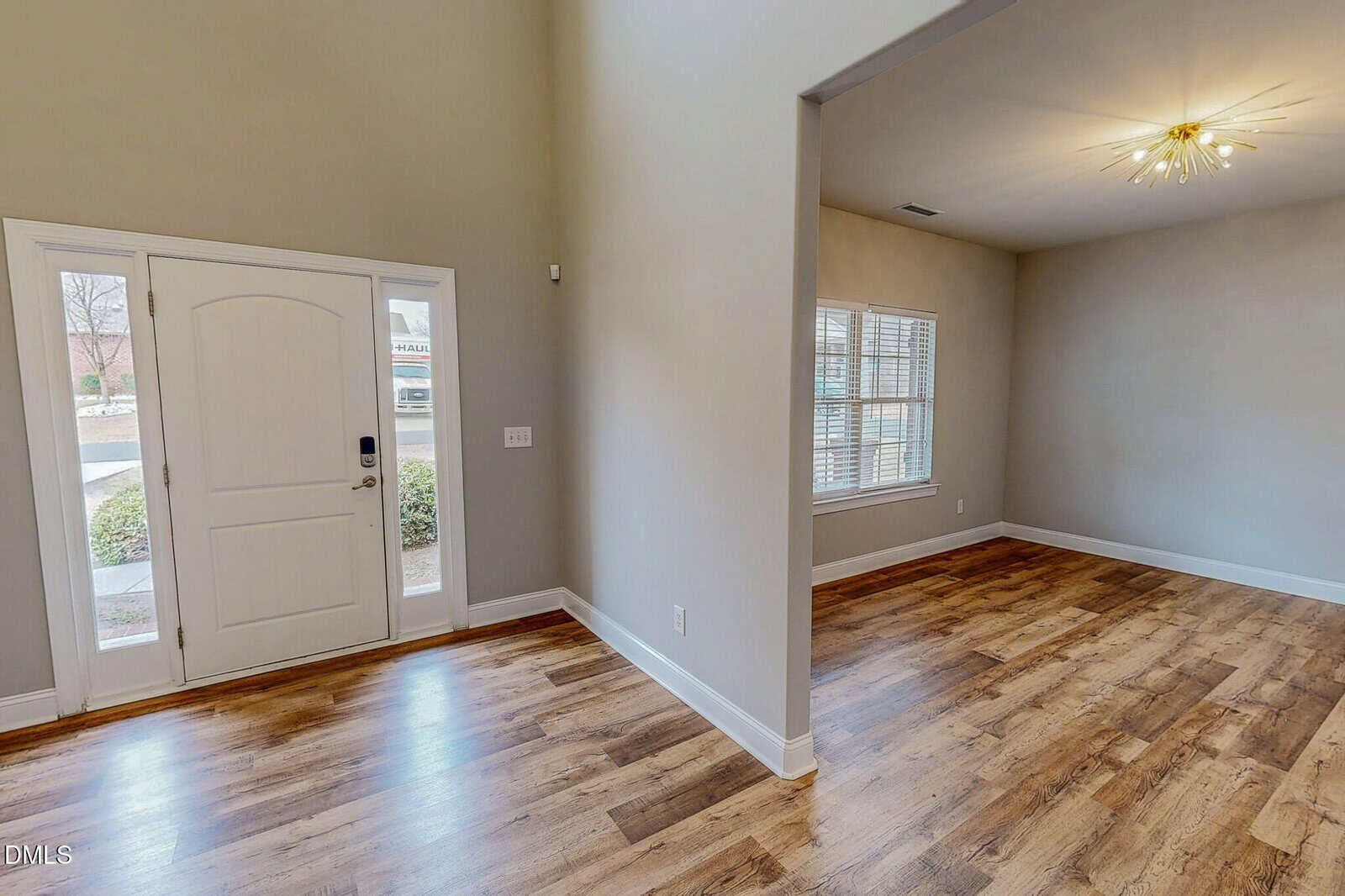 The image shows a home interior with an entryway and an adjacent room. The space has light beige walls, wood-style flooring, a large window, and a modern ceiling light fixture. A white front door is visible.