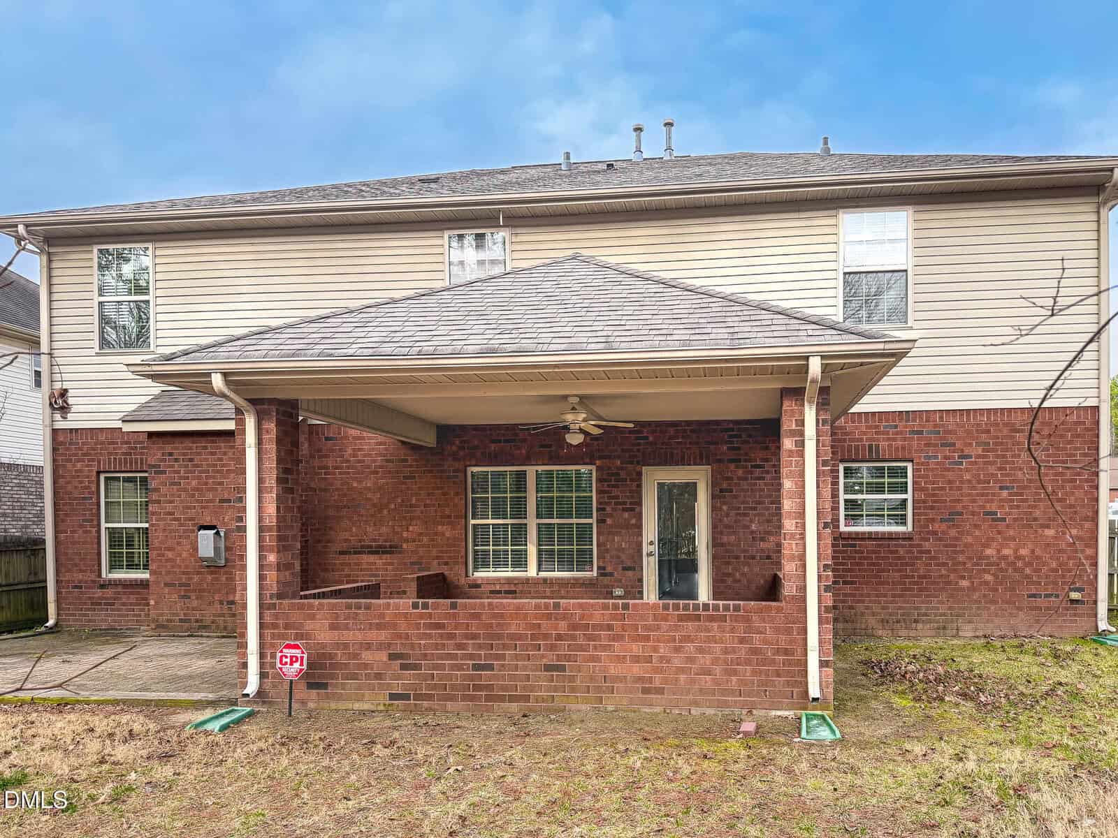 A two-story house with a covered back patio. The house exterior is partly red brick and partly beige siding. The patio has a ceiling fan, windows, a glass door, and a small yard with some grass.