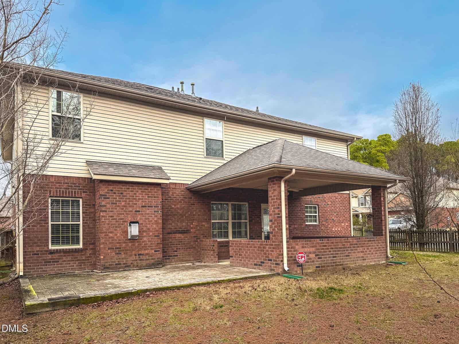 Two-story house with beige siding on the upper level and red brick on the lower level. There is a covered patio area with a concrete floor, surrounded by a bare yard and a few trees.