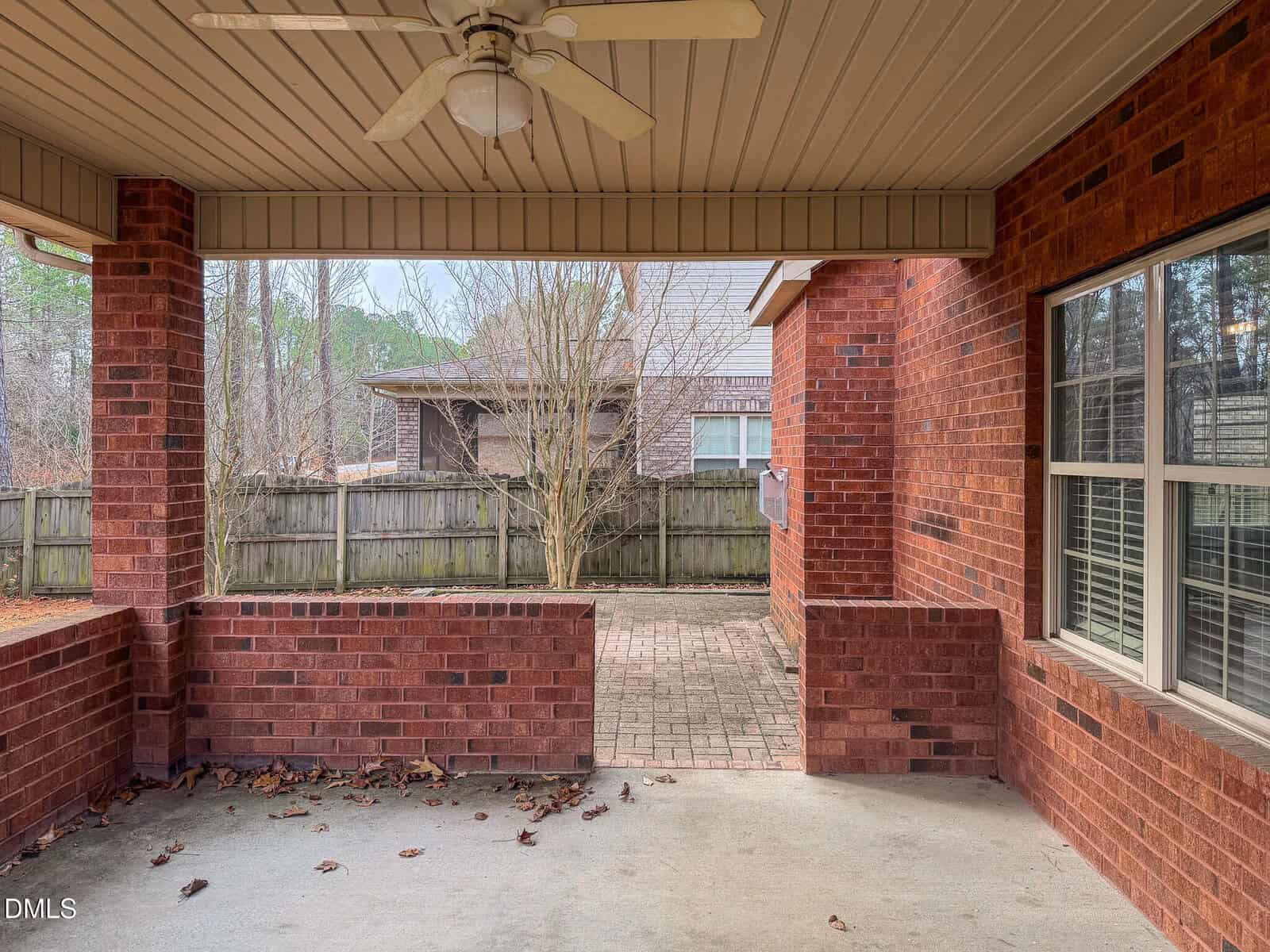 Covered brick patio with a ceiling fan, bordered by a partial brick wall. The patio overlooks a fenced backyard with bare trees and a neighboring brick house in the background. Leaves are scattered on the ground.