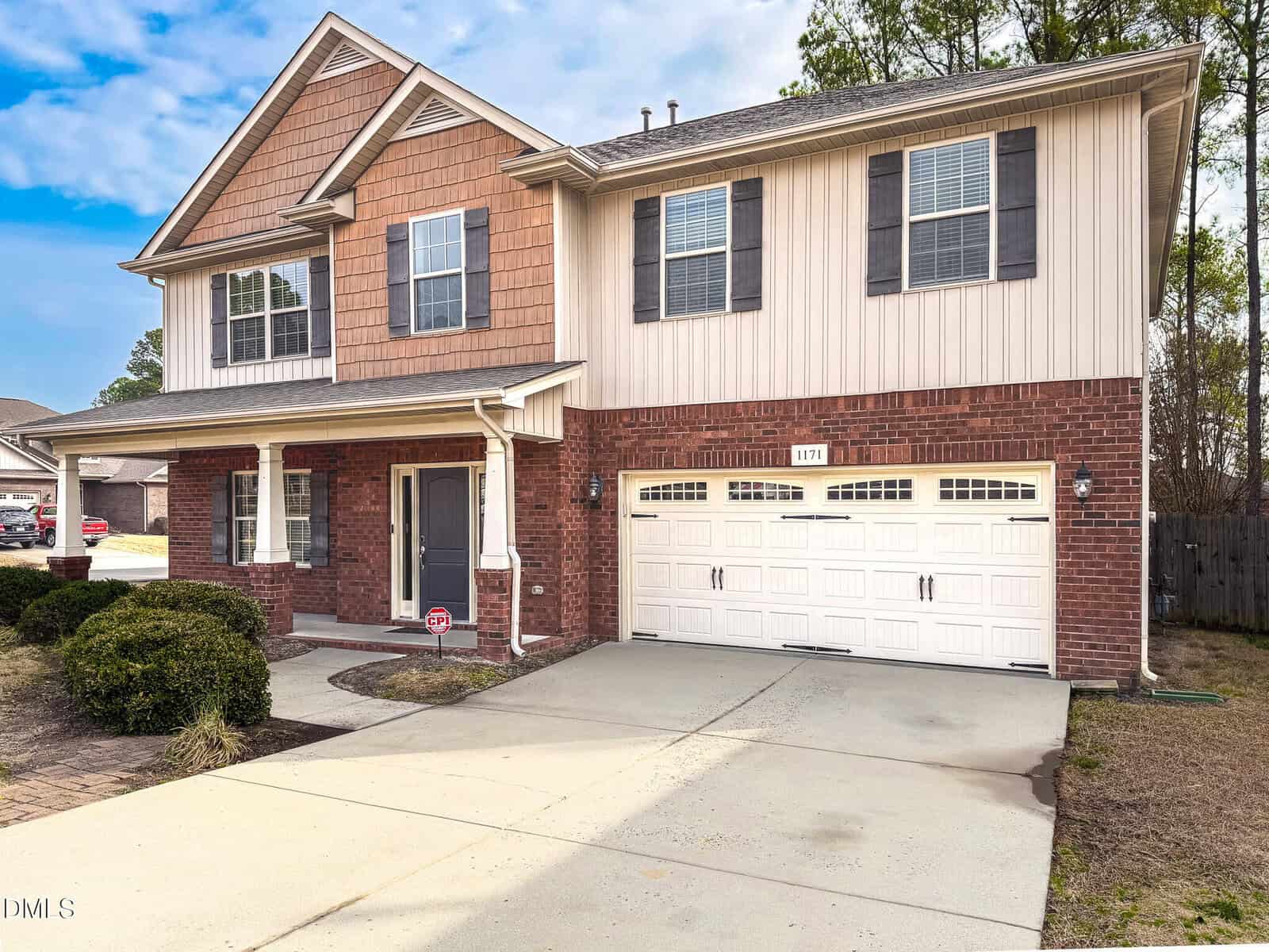 A two-story house with a brick and siding exterior, covered front porch, black front door, and a two-car white garage. The driveway is concrete, and there are bushes in the front yard.