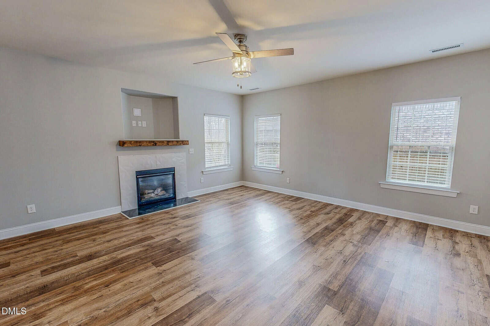 Spacious empty living room with light brown wood flooring, light gray walls, three windows, a ceiling fan with light, and a corner fireplace with a white mantel and built-in shelves above.