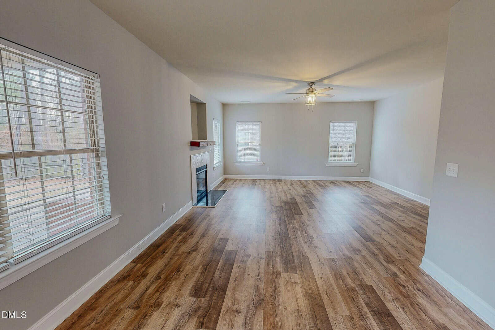 Spacious empty living room with wood flooring, neutral walls, ceiling fan, large windows, and a fireplace with a white mantle. Natural light brightens the space, giving it an open and inviting feel.