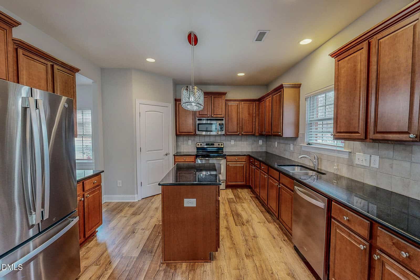 Modern kitchen with wood cabinets, stainless steel appliances, center island, tile backsplash, black countertops, wood floor, and windows letting in natural light. A round pendant light hangs above the island.