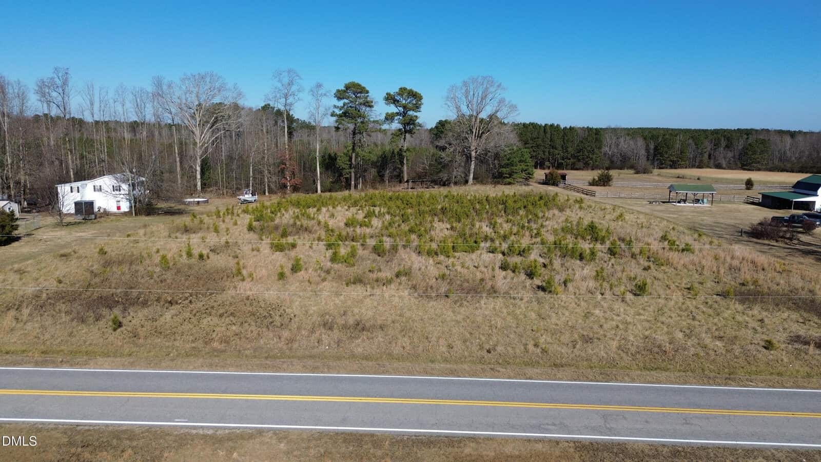 A grassy, undeveloped lot with small shrubs and trees sits beside 0 White Level Road in Louisburg, next to a white house near tree-lined woods; a green-roofed structure is visible on the right beneath a clear blue sky.
