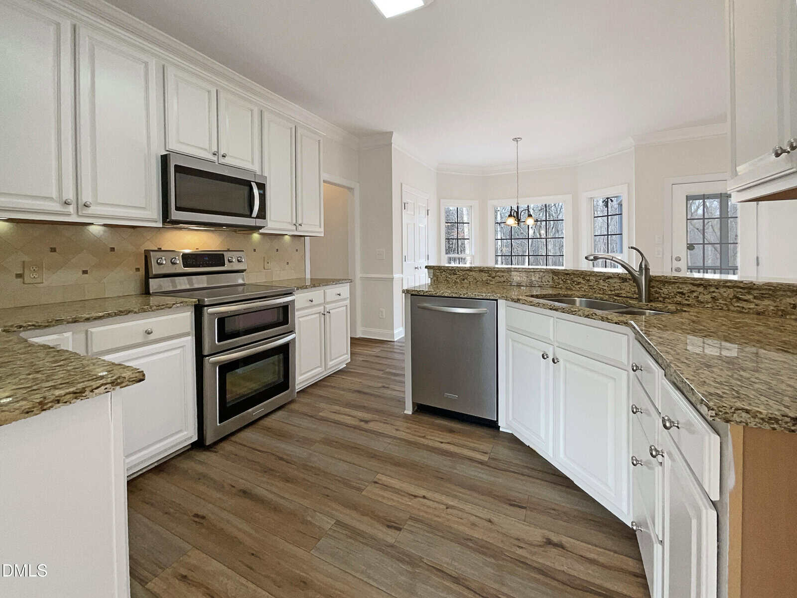 A spacious kitchen with white cabinets, granite countertops, stainless steel appliances, and wood-style flooring. Large windows and a glass door provide natural light and views of trees outside.
