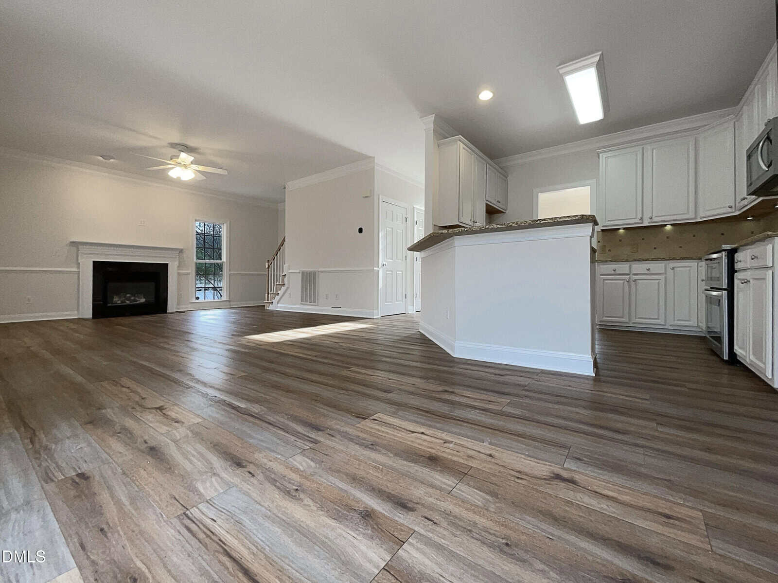 Spacious open-concept living area with wood-style flooring, white kitchen cabinets, granite countertops, a fireplace, ceiling fan, and staircase leading to an upper floor. Natural light fills the room through large windows.
