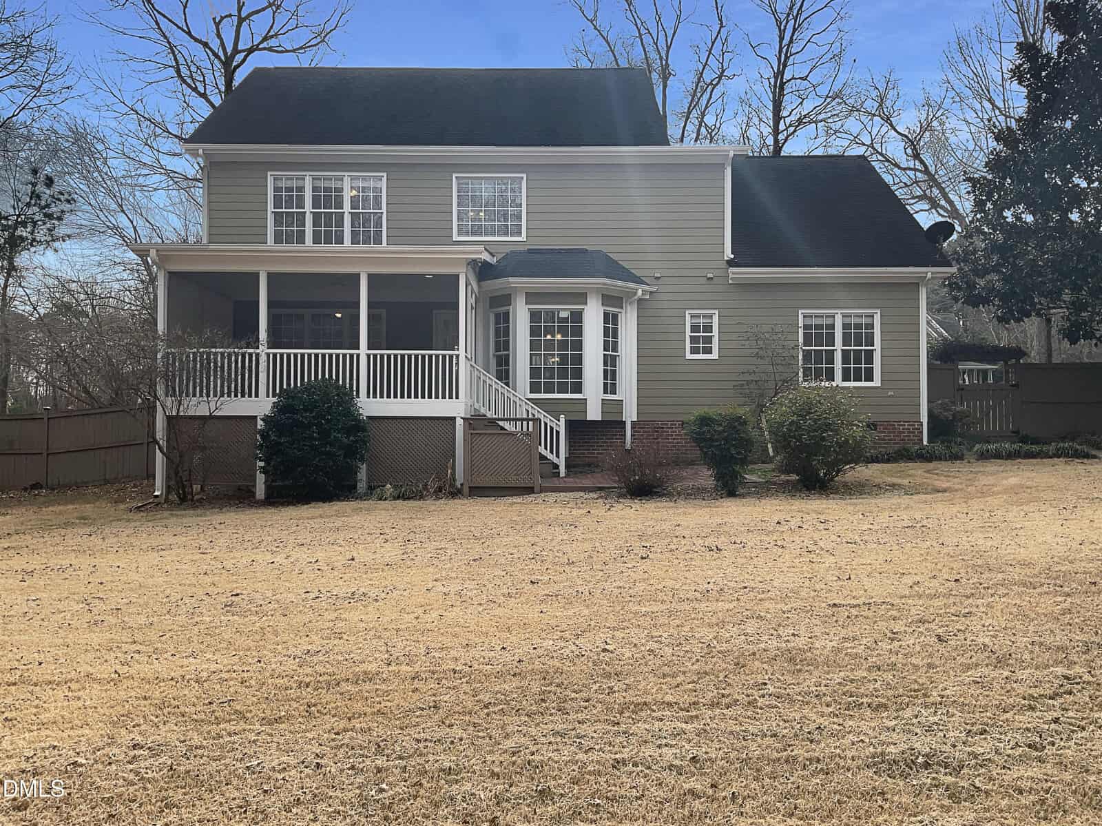Two-story house with beige siding, white trim, and a front porch with steps leading to a dry, yellowed lawn. There are a few bushes near the house and leafless trees in the background under a clear blue sky.