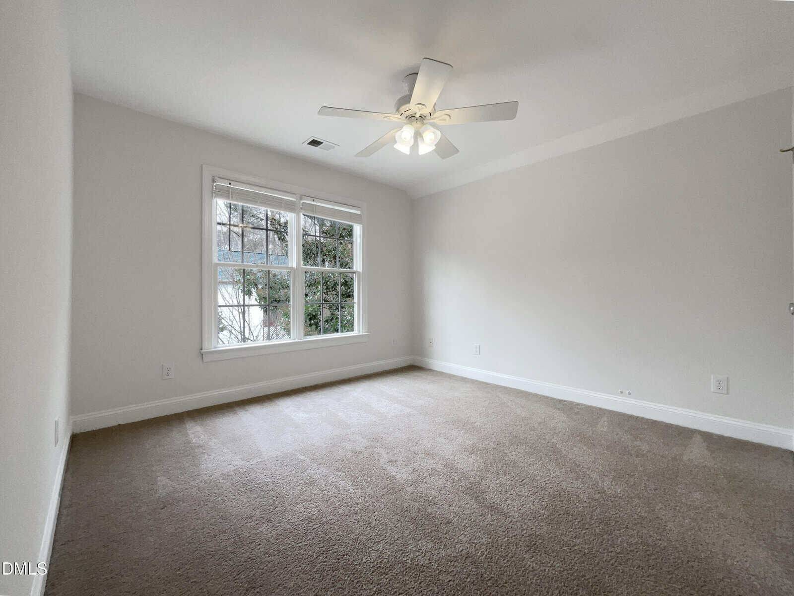 A bright, empty room with beige carpet, off-white walls, a ceiling fan with lights, and a large window letting in natural light. Greenery is visible through the window.