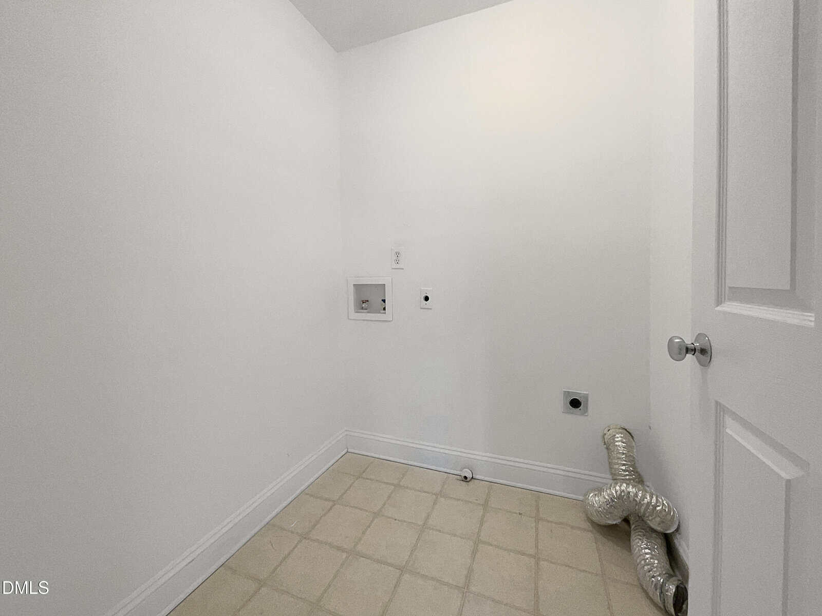 A small, empty laundry room with white walls, beige tiled floor, washer and dryer hookups, and a flexible metal vent hose lying on the floor in the corner.