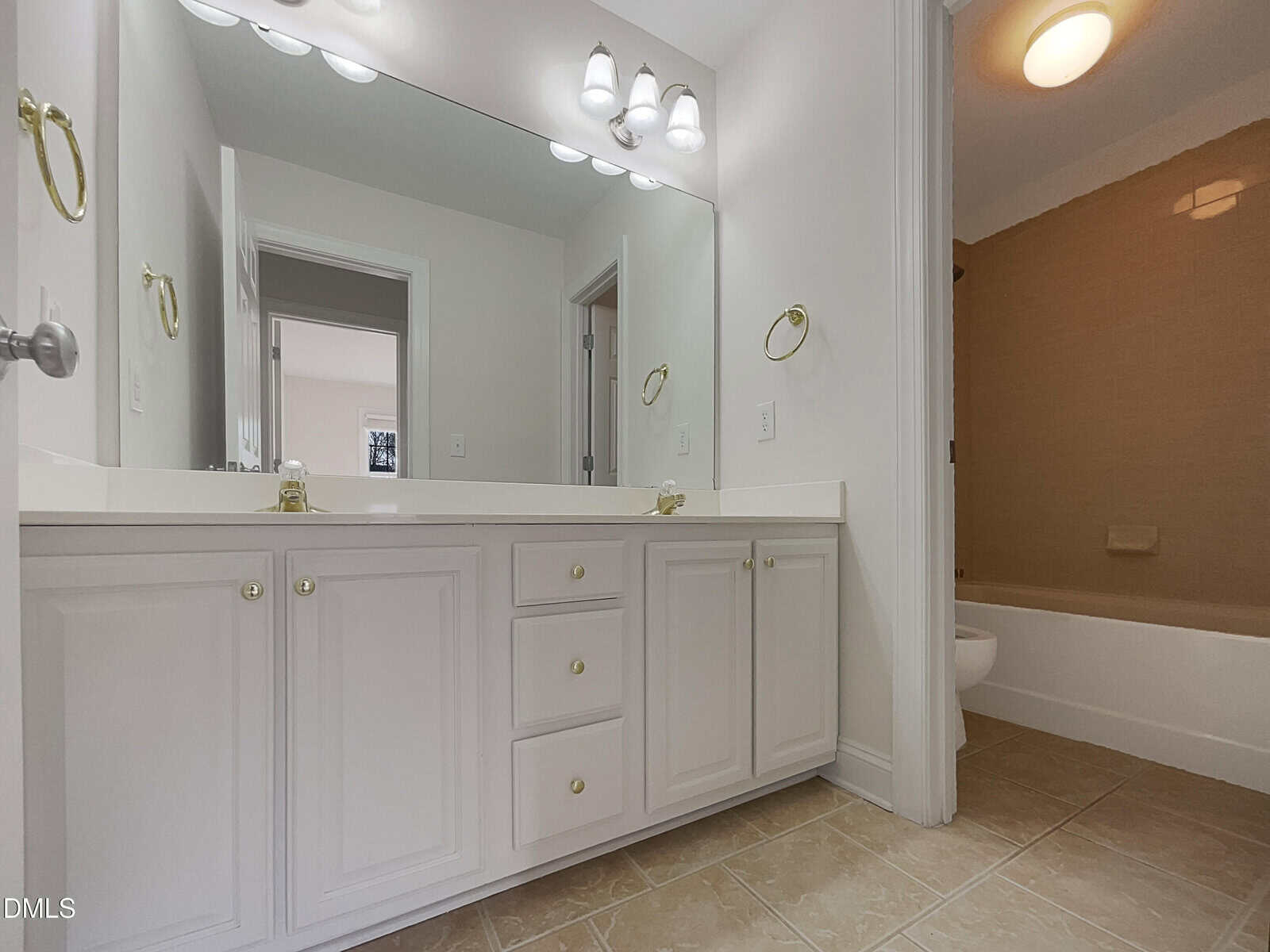 A bathroom with a large mirror above a white double-sink vanity with gold hardware, beige tiled floor, and a bathtub with tan tile walls in the background. The walls are light-colored and the room is well-lit.