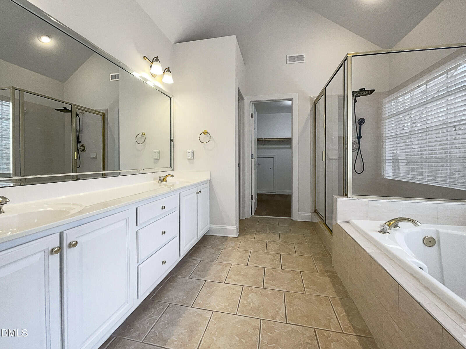 Spacious bathroom with double sinks, large mirrors, white cabinets, tan tile floor, a glass-enclosed shower, and a bathtub. The room is well-lit with natural light from a window and has vaulted ceilings.