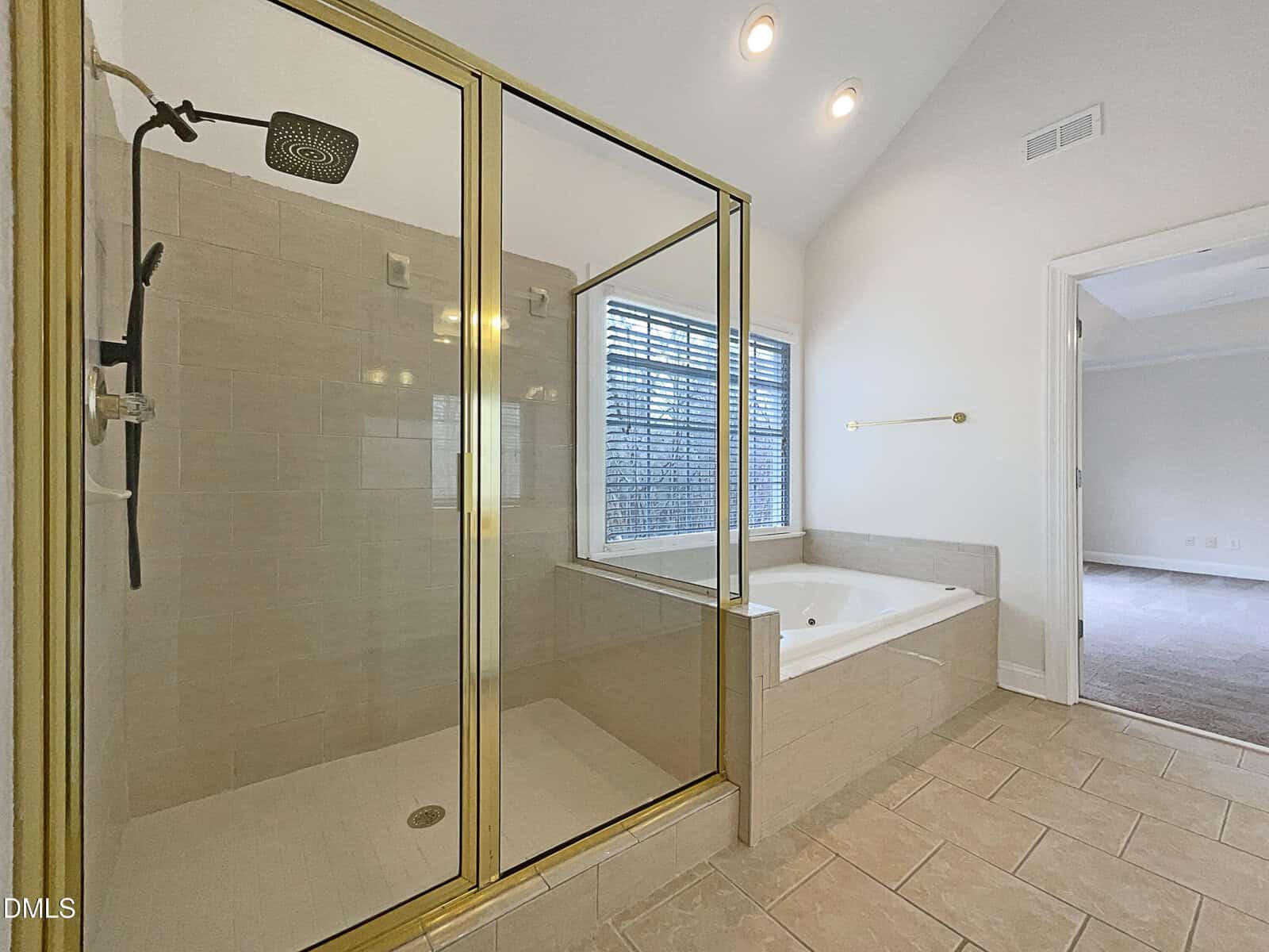 Modern bathroom with a large glass-enclosed shower, a soaking tub beside a window, beige tile flooring, and white walls. The space leads into an adjacent carpeted bedroom.