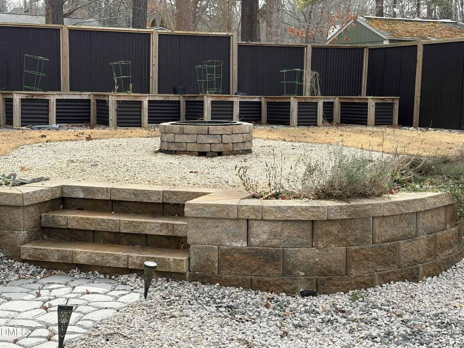 A backyard with a circular stone fire pit on gravel, surrounded by a low curved stone retaining wall, steps, garden beds with metal plant supports, and a black privacy fence.