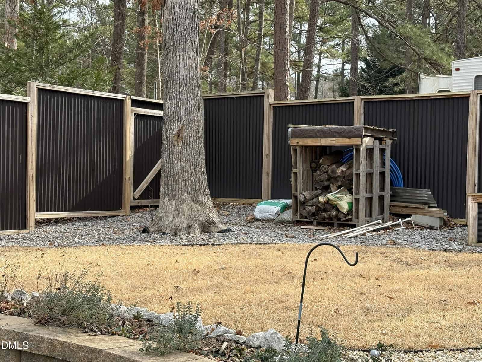 A backyard with a tall black fence, a large tree, a woodpile under a small shelter, garden supplies, and a patch of dry grass in the foreground. Pines and other trees are visible beyond the fence.