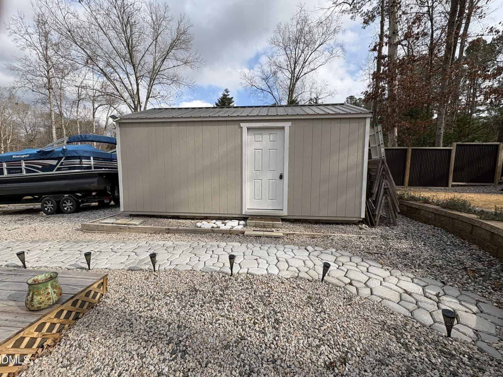 A beige shed with a white door and metal roof sits on a gravel lot. A boat on a trailer is parked to the left, and trees without leaves are in the background under a partly cloudy sky.
