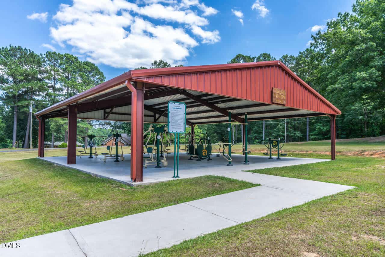 A covered outdoor gym with exercise equipment sits on a concrete pad, surrounded by grass and trees under a partly cloudy sky. A sign with information stands near the entrance.