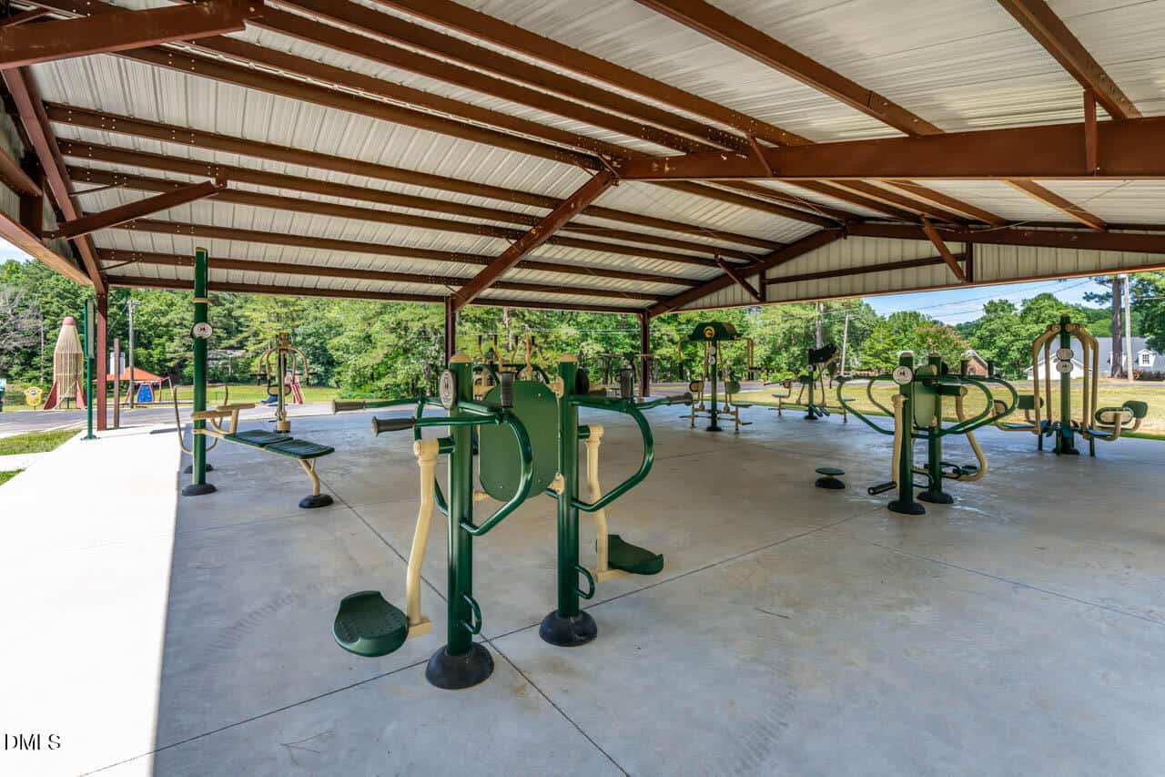 Outdoor gym under a metal roof with various green exercise equipment on a concrete floor, surrounded by trees and grass on a sunny day.