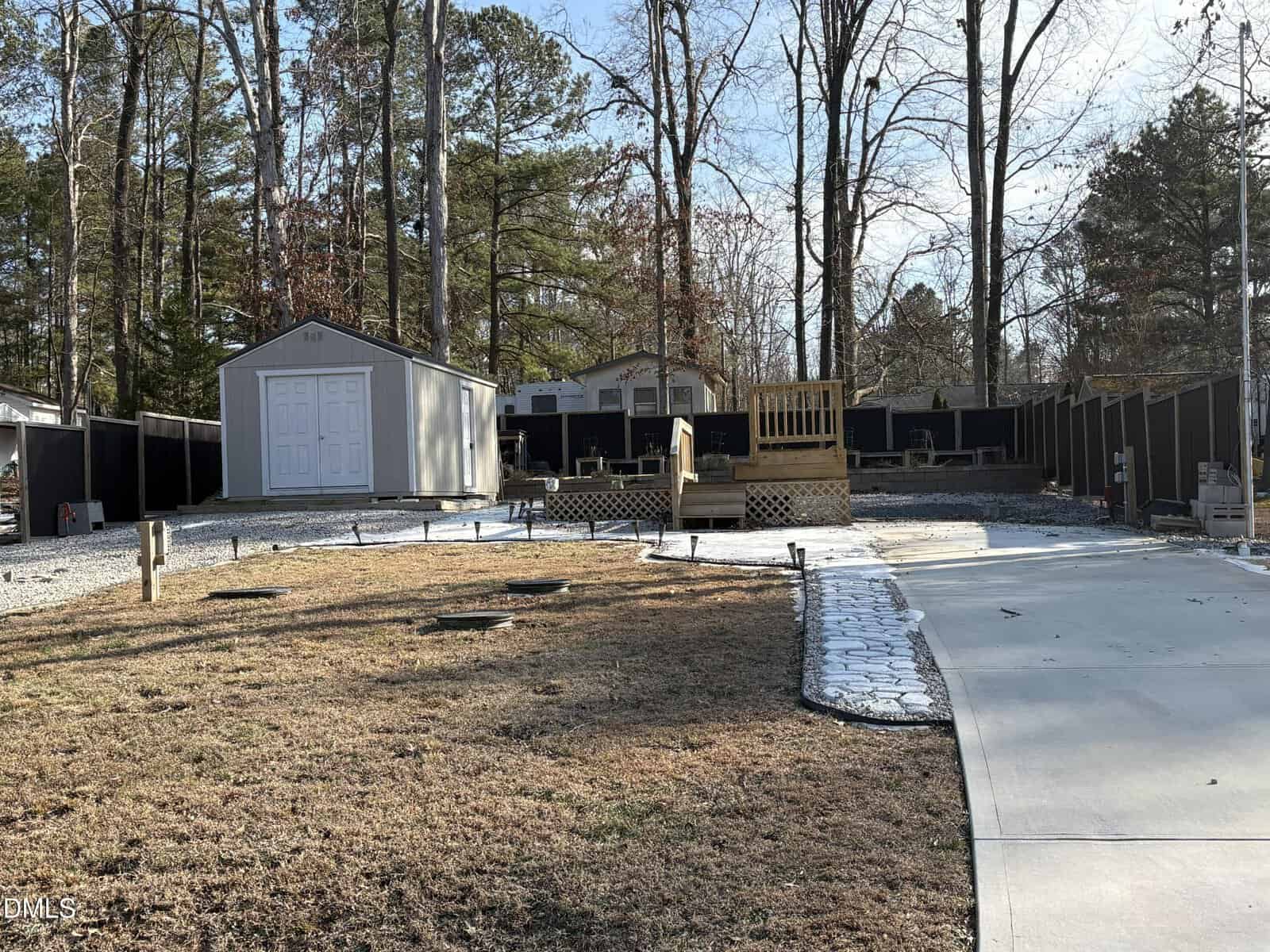 A backyard with a shed on the left, a small wooden deck in the center, a patch of grass, a concrete walkway, and trees in the background. A black fence runs along the perimeter.