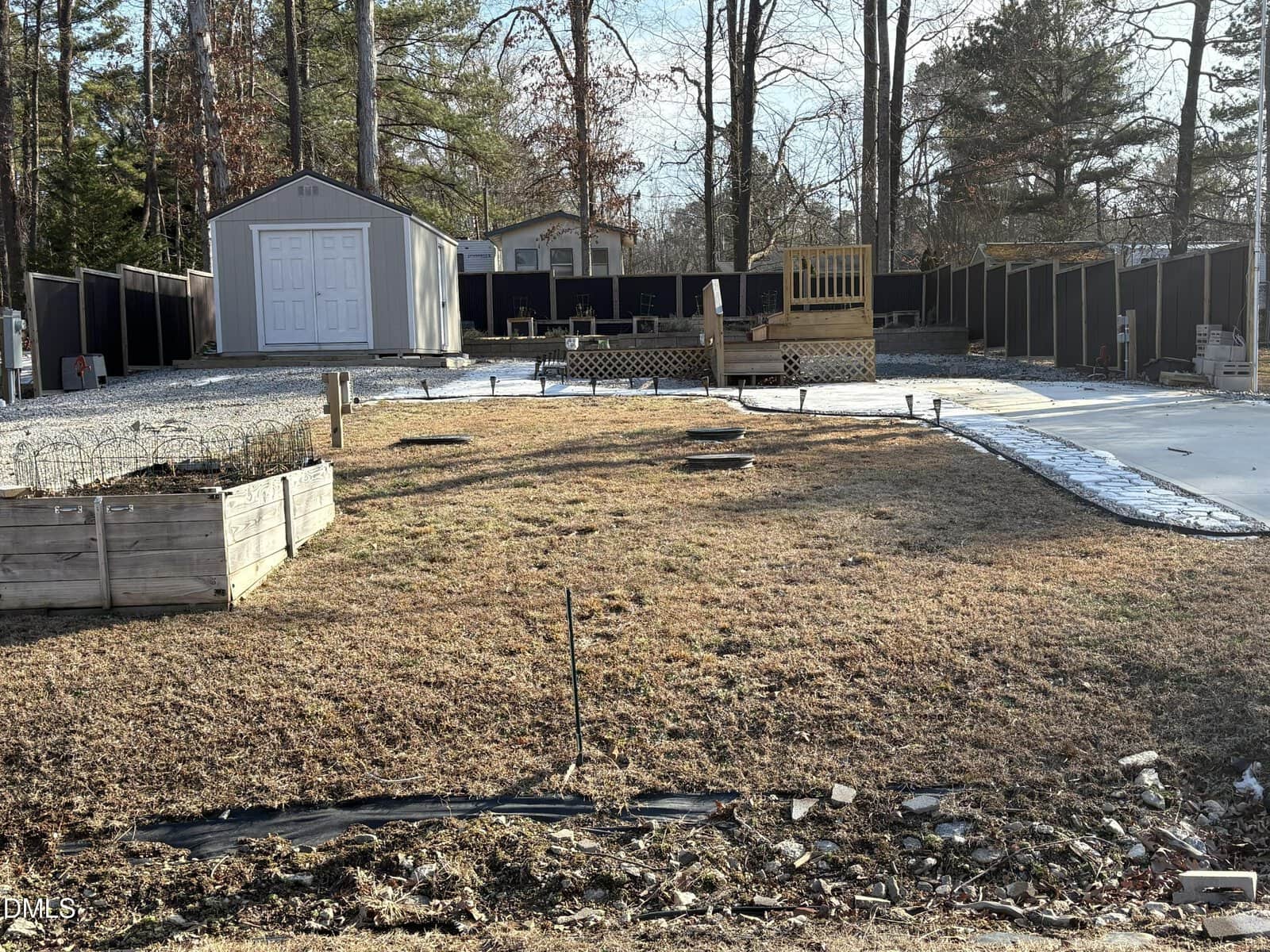A backyard with brown grass, a raised garden bed on the left, a storage shed in the back left corner, a wooden deck in the center, and a black privacy fence surrounding the area. Trees are visible in the background.