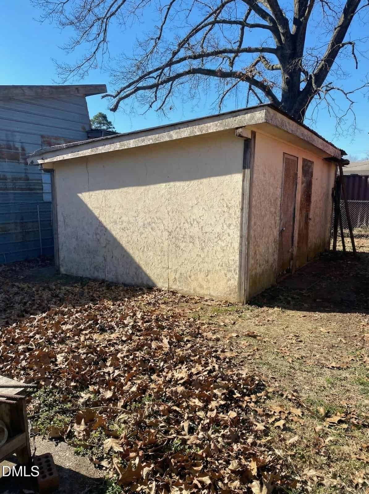 A small, weathered shed with a slanted roof stands in a backyard. Fallen leaves cover the ground, and a bare tree towers behind the shed under a clear sky.