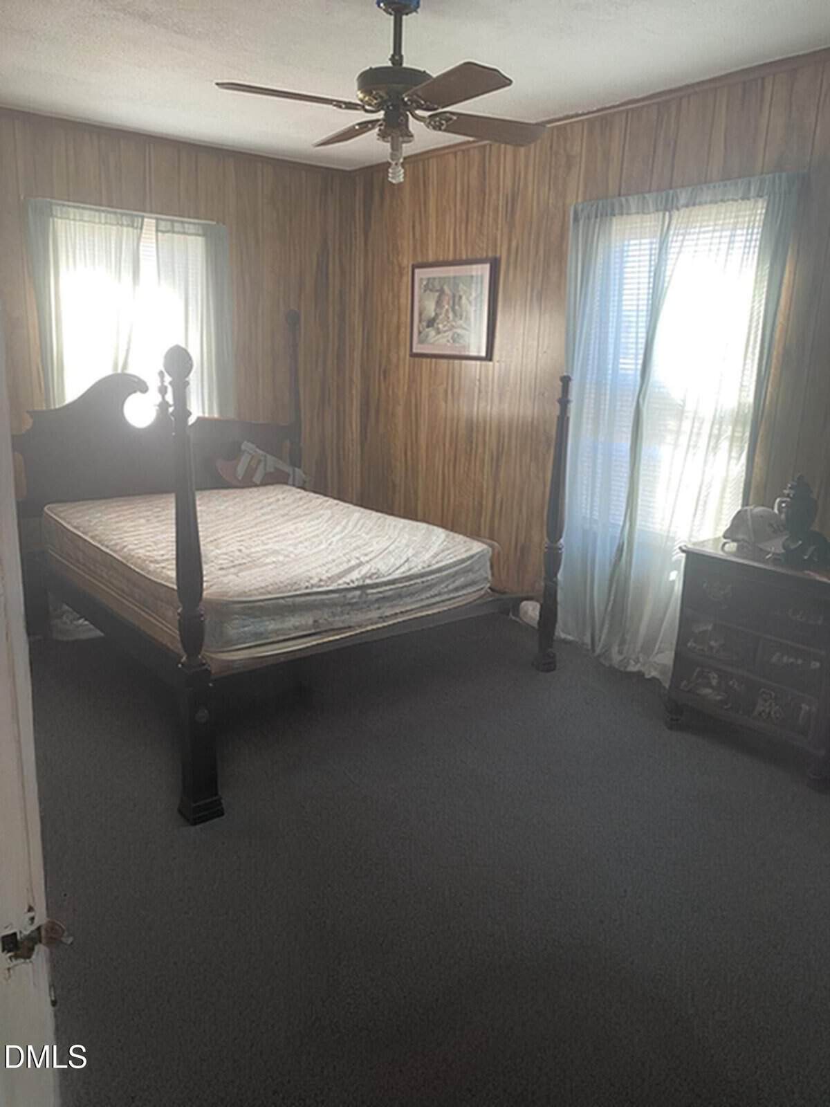 A bedroom with wood-paneled walls, a ceiling fan, a four-poster bed without bedding, two windows with sheer curtains, a framed picture on the wall, and a dark wooden dresser with items on top.