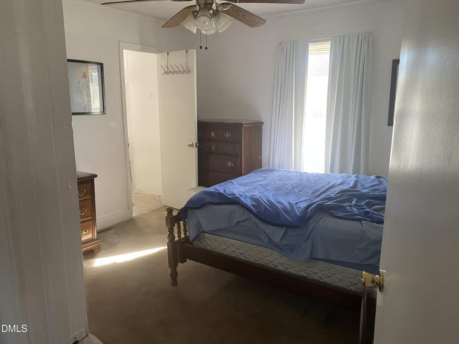 A small bedroom with a wooden bed frame, blue bedding, chest of drawers, and two windows with white curtains. A ceiling fan is above, and an open door reveals part of a bathroom.