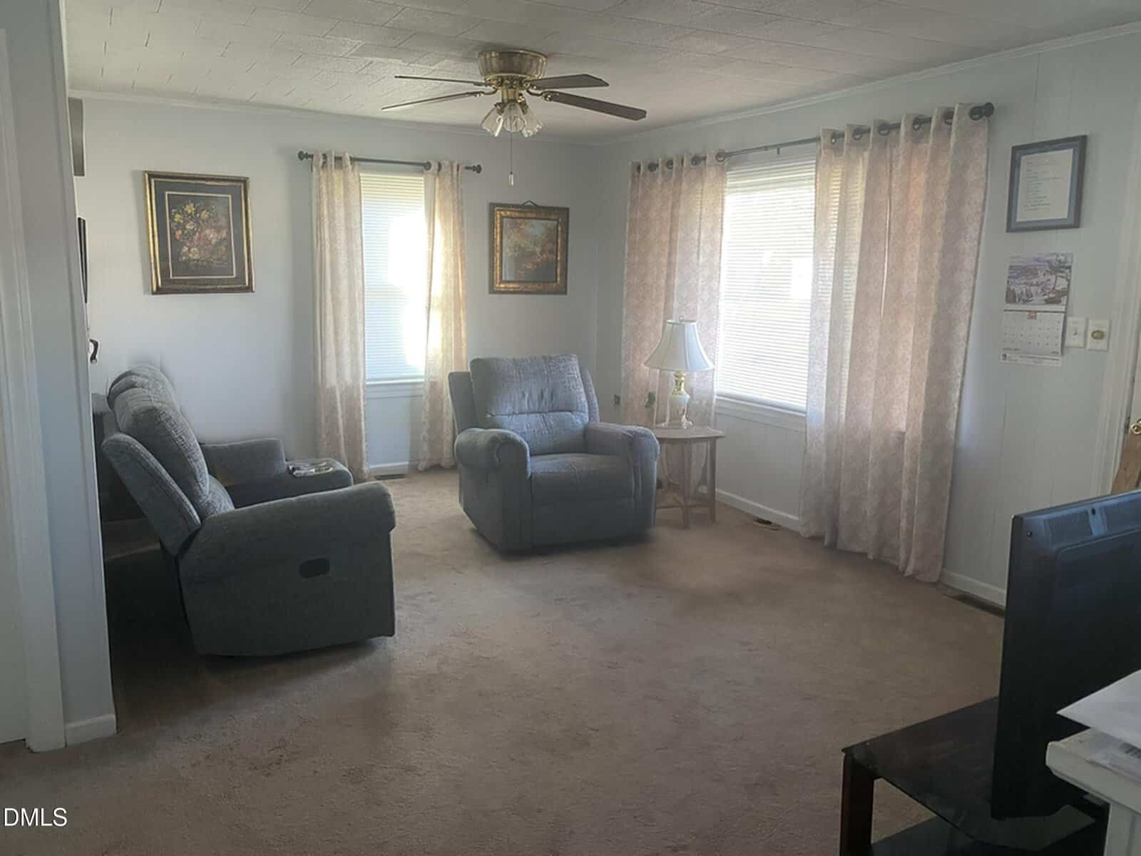 A living room with light-colored walls, beige carpet, two gray recliner chairs, a side table with a lamp, framed artwork, two windows with curtains, and a ceiling fan with lights. A TV is visible in the foreground.