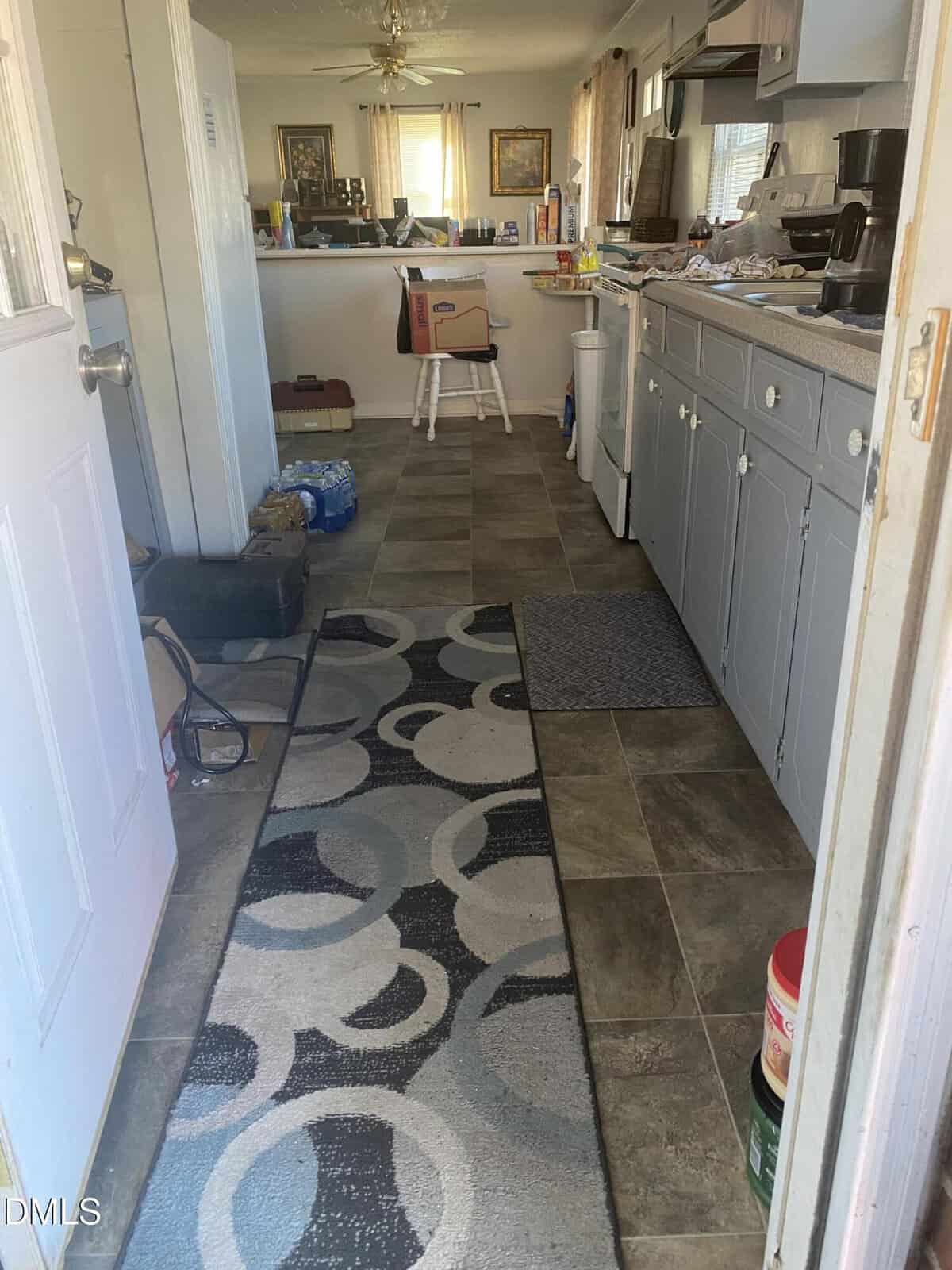 A view of a narrow kitchen with gray cabinets, countertop appliances, and a patterned rug on a tiled floor. Boxes, water bottles, and various items are scattered throughout the space. Sunlight enters from windows.