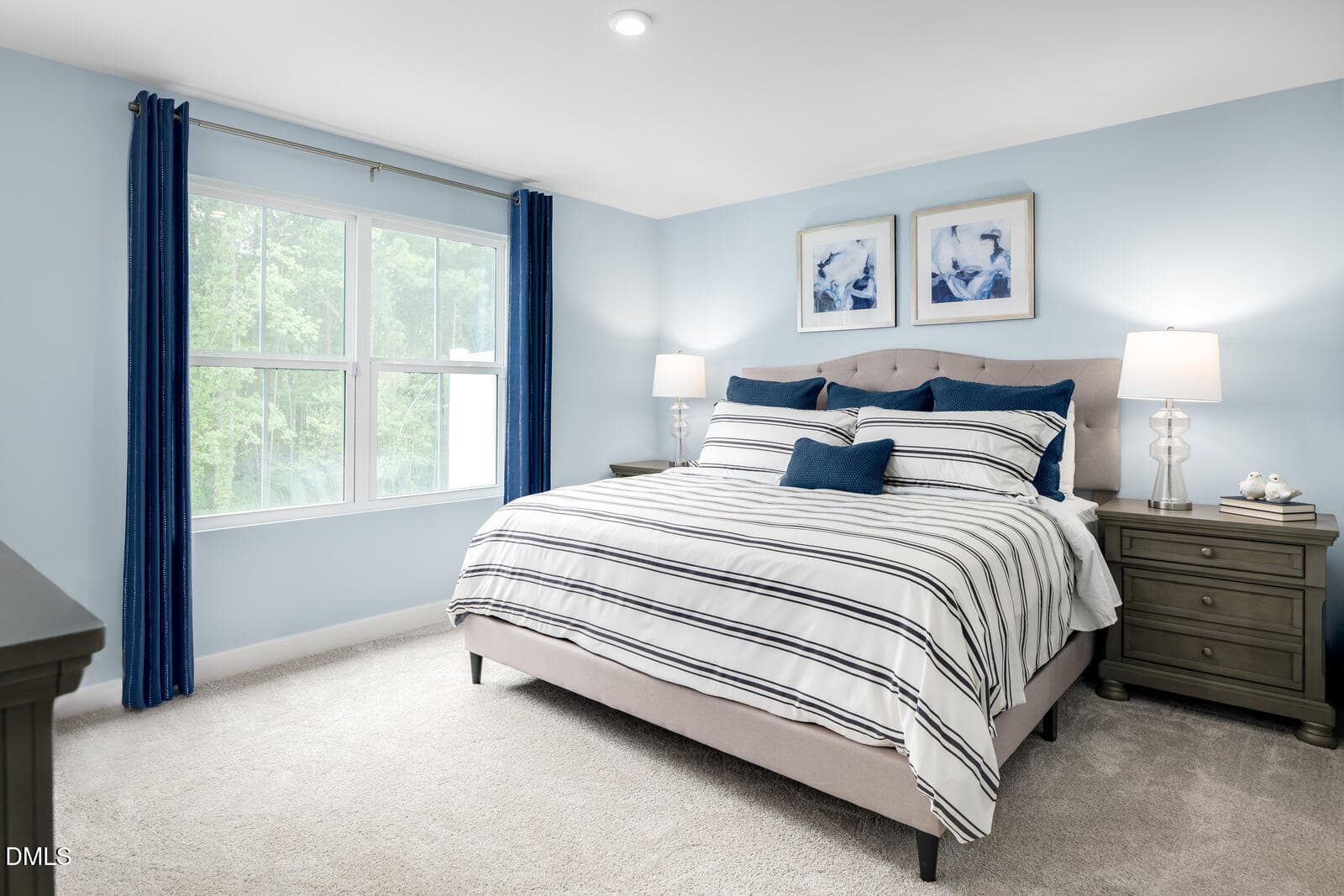 Bright bedroom with a large bed, striped bedding, blue accent pillows, two bedside tables with lamps, blue curtains, and two abstract artworks on a light blue wall. Large window lets in natural light.