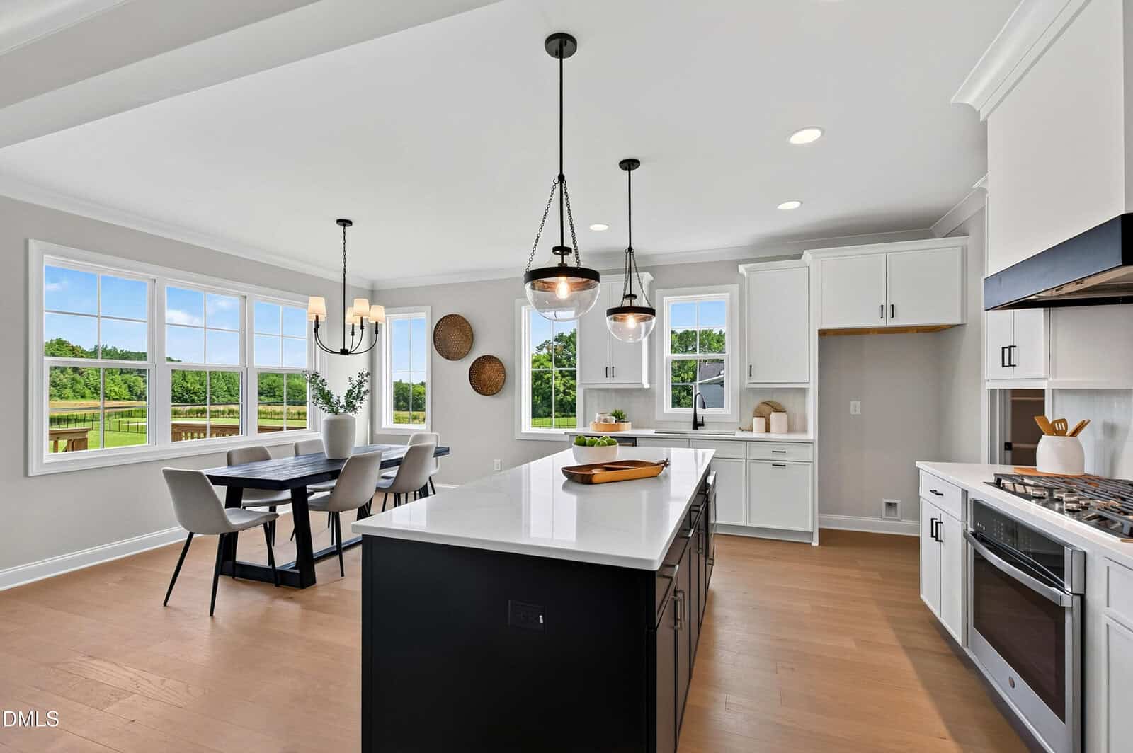 Bright modern kitchen with white cabinets, a large island with pendant lights, dining area with six chairs, and large windows overlooking a green outdoor landscape.
