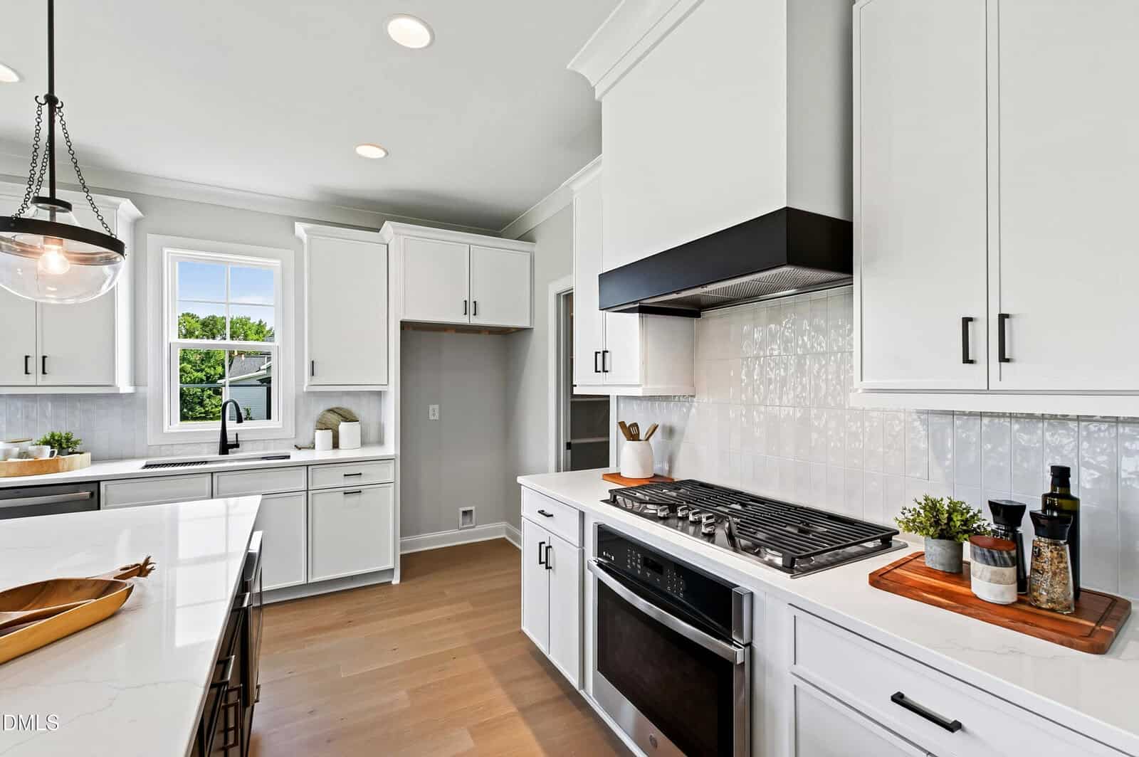 Modern kitchen with white cabinets, a gas stove with a black hood, built-in oven, marble countertops, island with sink, wood floors, and decorative items. Window lets in natural light; pantry area is visible in the background.