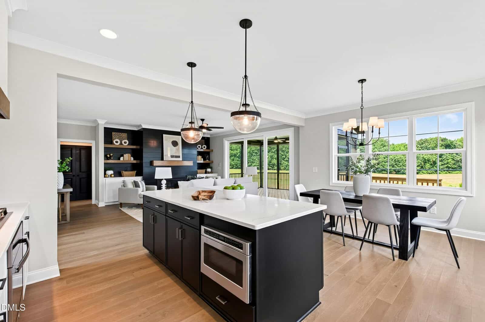 Modern kitchen with a black island, pendant lights, white countertops, wood floors, dining area with large windows, and living space in the background. Bright, open layout with outdoor views.