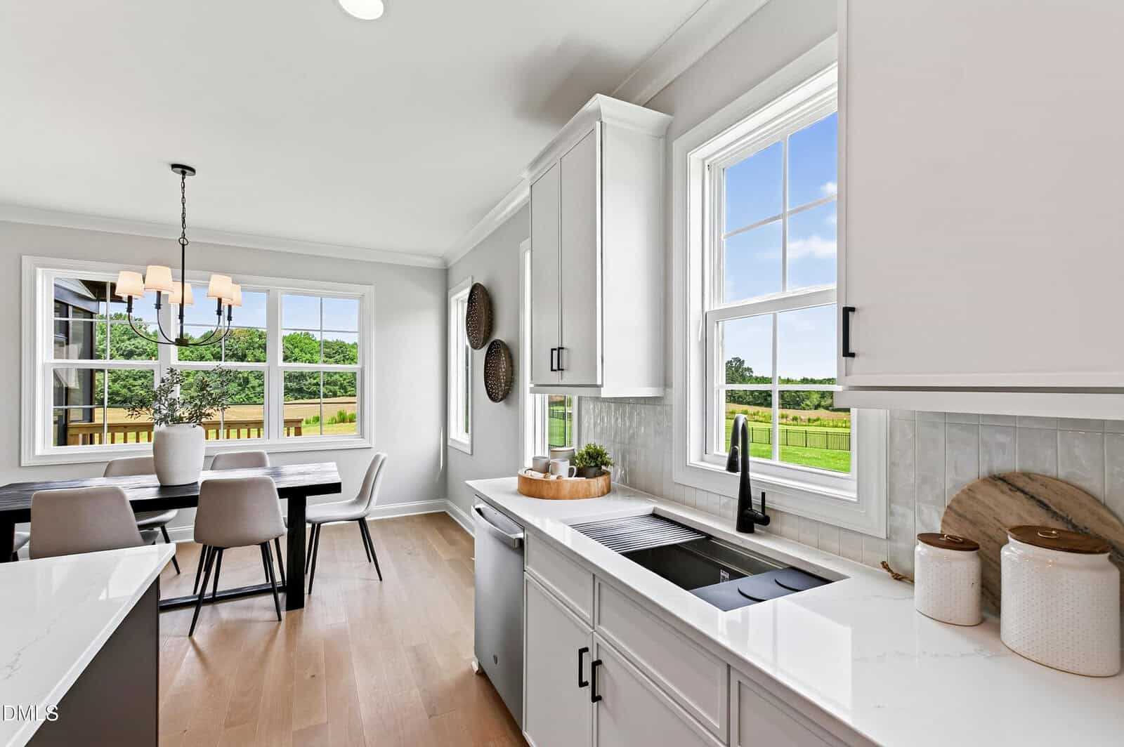 Bright modern kitchen with white cabinets, black fixtures, and large windows overlooking a green backyard. Dining area with a black table and six chairs is visible, illuminated by a chandelier.