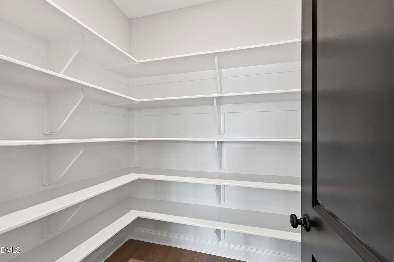 Empty walk-in pantry with white shelves on three walls, beige walls, and a dark door with a round handle. The floor is wooden, and the shelving is arranged in an L-shape with multiple levels.
