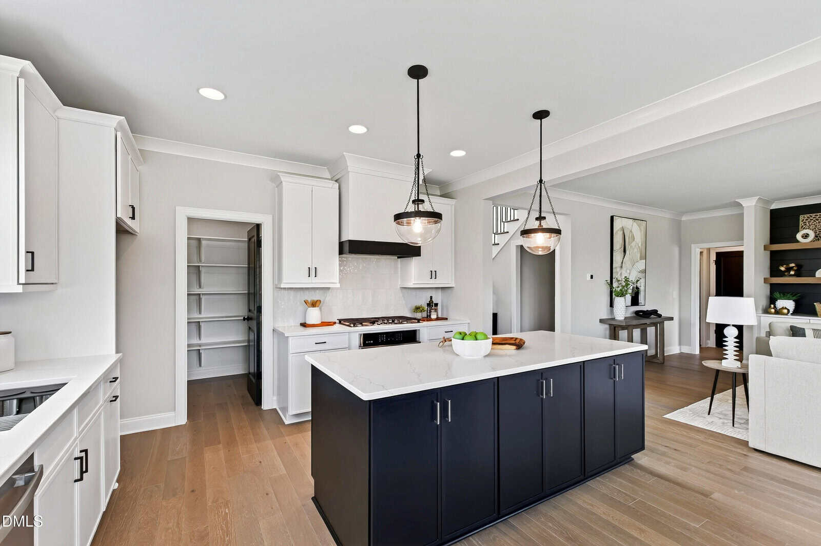 Modern kitchen with white cabinets, a black island with a white countertop, pendant lights, and wood flooring. Open doorway leads to a pantry, and a living area is partially visible in the background.