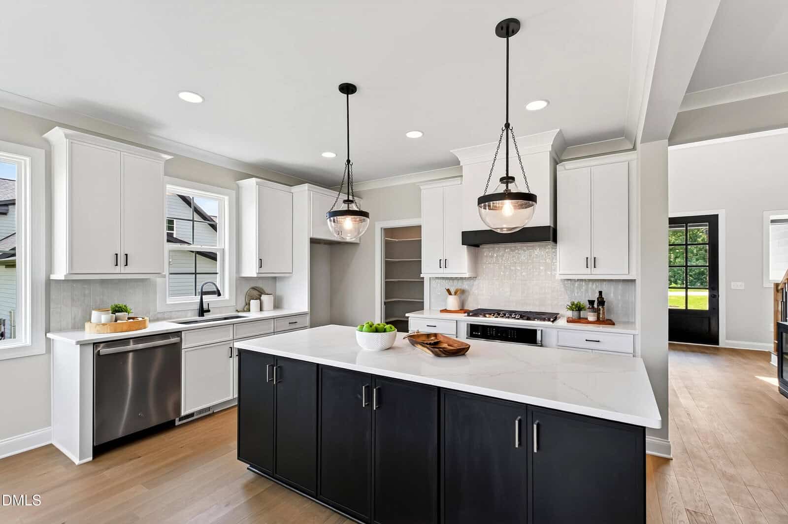 Modern kitchen with white upper cabinets, black island, white countertops, stainless steel appliances, pendant lights, wood floors, a large farmhouse sink, and windows providing natural light.