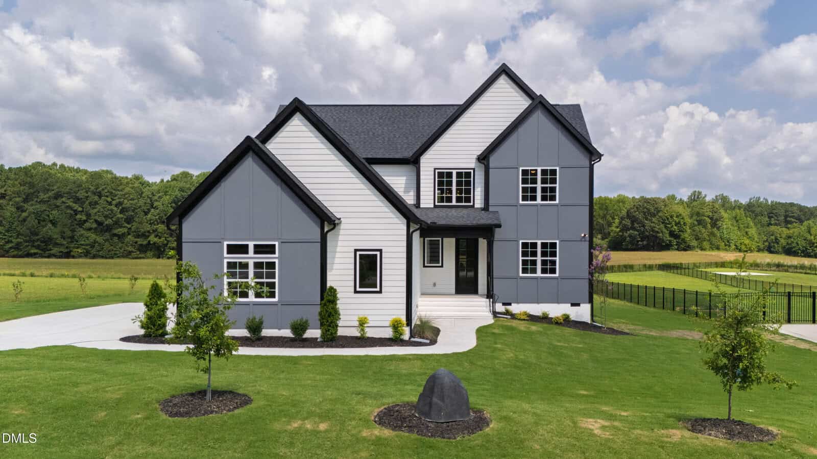 Two-story modern farmhouse with a mix of gray and white siding, black trim, a dark shingle roof, and a large front lawn with young trees and shrubs. A stone sits in the center of the grass near the driveway.