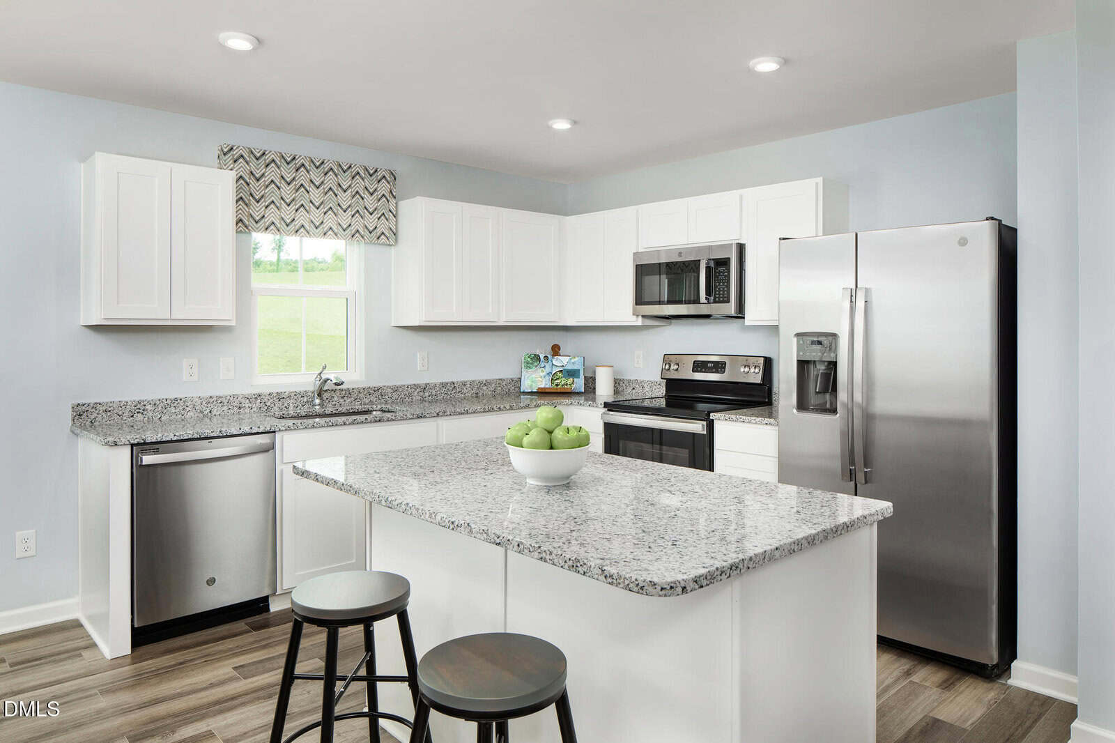 A modern kitchen with white cabinets, stainless steel appliances, a granite island with two stools, a bowl of green apples, and a window with a patterned valance. The flooring is light wood.